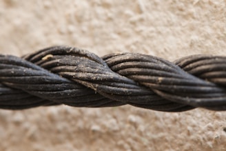 A close-up view of a thick, black rope composed of multiple twisted strands. The rope is slightly dusty, indicating it might have been outdoors. The background is a textured, beige surface that appears blurred in comparison to the sharply focused rope.
