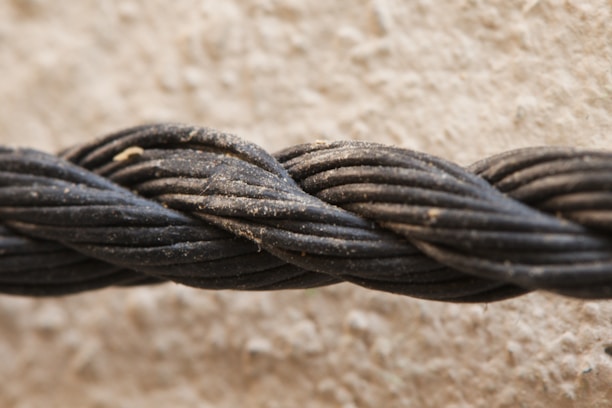 A close-up view of a thick, black rope composed of multiple twisted strands. The rope is slightly dusty, indicating it might have been outdoors. The background is a textured, beige surface that appears blurred in comparison to the sharply focused rope.