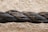 A close-up view of a thick, black rope composed of multiple twisted strands. The rope is slightly dusty, indicating it might have been outdoors. The background is a textured, beige surface that appears blurred in comparison to the sharply focused rope.
