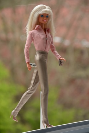 A blonde fashion doll is poised elegantly, wearing a pink blouse and beige high-waisted pants. It holds a small object in one hand. The background is blurred with hints of greenery and a reddish hue.