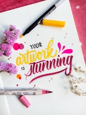 A colorful, artistic arrangement features the phrase 'Your artwork is stunning' in vibrant lettering. The words are surrounded by markers, one with a yellow tip and another with a pink tip. Accents include small floral elements, both pink flowers and white delicate sprigs, adding a decorative touch to the scene.
