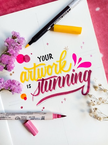 A colorful, artistic arrangement features the phrase 'Your artwork is stunning' in vibrant lettering. The words are surrounded by markers, one with a yellow tip and another with a pink tip. Accents include small floral elements, both pink flowers and white delicate sprigs, adding a decorative touch to the scene.