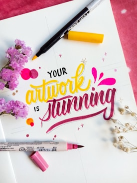 A colorful, artistic arrangement features the phrase 'Your artwork is stunning' in vibrant lettering. The words are surrounded by markers, one with a yellow tip and another with a pink tip. Accents include small floral elements, both pink flowers and white delicate sprigs, adding a decorative touch to the scene.