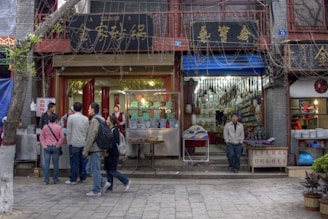 A bustling street scene with several people gathered outside small shops. The shops have signs with Chinese characters, and one of the shops appears to be selling food, as indicated by the people queuing and the vendor inside. A man stands casually in front of a neighboring shop filled with various items hanging from the ceiling.