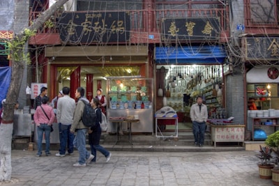 A bustling street scene with several people gathered outside small shops. The shops have signs with Chinese characters, and one of the shops appears to be selling food, as indicated by the people queuing and the vendor inside. A man stands casually in front of a neighboring shop filled with various items hanging from the ceiling.