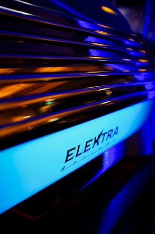 An illuminated blue strip with the word 'ELEKTRA' written on it, likely part of a machine or device. The surrounding area is dark with reflections of warm orange and purple hues on metallic ridges.