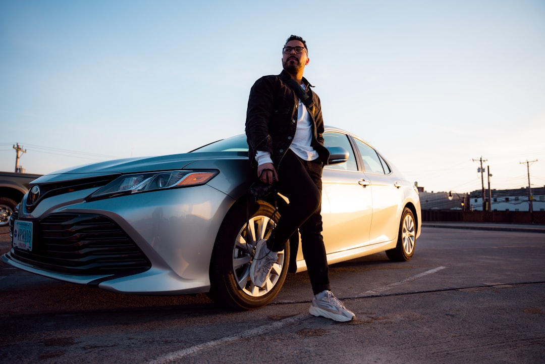 man in black jacket and blue denim jeans leaning on silver sedan