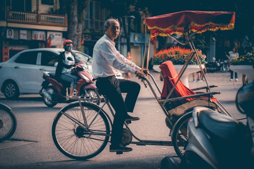A person wearing a white shirt is riding a traditional rickshaw on a city street. The rickshaw has a red canopy with ornate yellow fringes, and a red seat cover. In the background, there is traffic including a person on a red scooter and a parked white car. The street is flanked by buildings and people walking by.