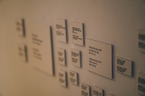 A series of small, white plaques are mounted on a wall. Each plaque features printed text and appears to be formatted as an informational display, possibly for an art exhibit. The focus is on one central plaque, with others slightly out of focus, creating a depth effect.