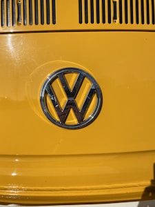 A close-up of a yellow vehicle's exterior showcasing the Volkswagen emblem in the center. The shiny metallic logo stands out against the bright yellow paint, with air vents visible at the top of the image.