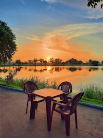 A serene lakeside view during sunset, featuring a tranquil body of water bordered by trees. In the foreground, a set of brown plastic chairs and a table are placed on a patio, suggesting a spot for relaxation and observation.