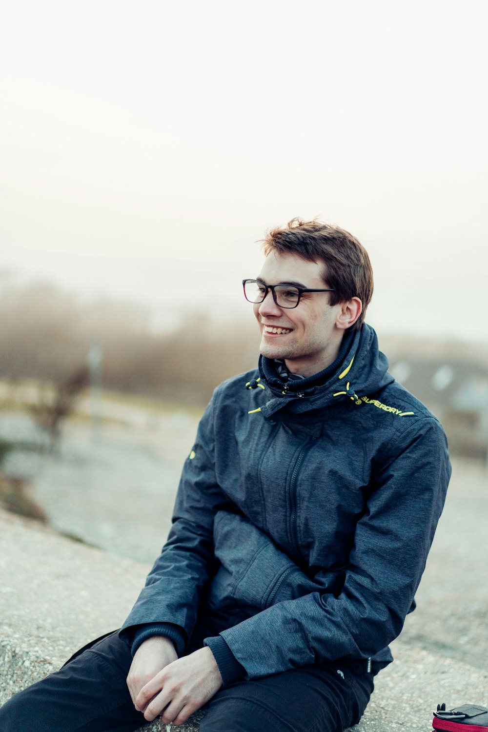 man in blue jacket and black framed eyeglasses sitting on gray concrete pavement during daytime