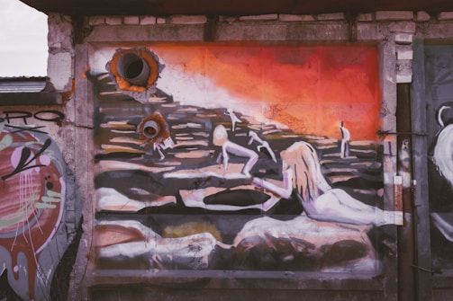 A mural depicting abstract human figures positioned against a background of warm, fiery hues. Two prominent pipe-like structures protrude from the wall, integrated into the artwork, creating a juxtaposition of industrial and artistic elements. The figures appear to interact with an undulating landscape, suggesting motion and activity.