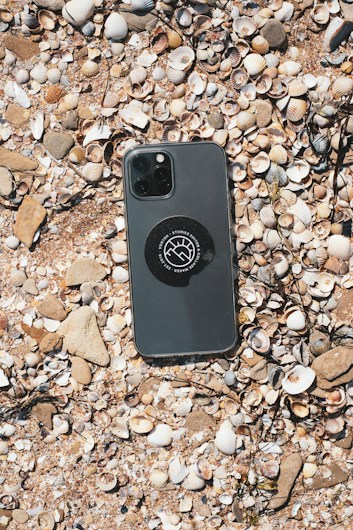 A smartphone rests on a surface covered with various seashells and small pebbles. The phone has a dark casing, with a circular sticker displaying a logo in the center. The shells vary in size and color, creating a textured background in natural tones.