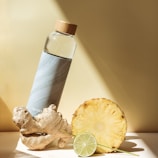 A stylish water bottle with a bamboo cap is placed next to a piece of ginger, a pineapple slice, and a lime slice, all on a light-colored surface. The scene is well-lit with shadows, creating a fresh and healthy vibe.