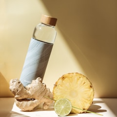 A stylish water bottle with a bamboo cap is placed next to a piece of ginger, a pineapple slice, and a lime slice, all on a light-colored surface. The scene is well-lit with shadows, creating a fresh and healthy vibe.