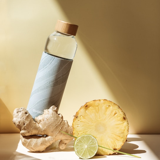A stylish water bottle with a bamboo cap is placed next to a piece of ginger, a pineapple slice, and a lime slice, all on a light-colored surface. The scene is well-lit with shadows, creating a fresh and healthy vibe.