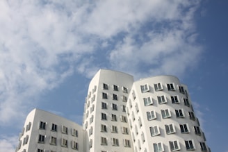 Curved modern white buildings with multiple windows reaching up towards a partly cloudy blue sky.