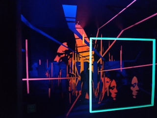 A vibrant, abstract artwork featuring neon outlines of human faces and geometric shapes. The composition includes intersecting lines and rectangles, creating a sense of depth and dimension against a dark background.