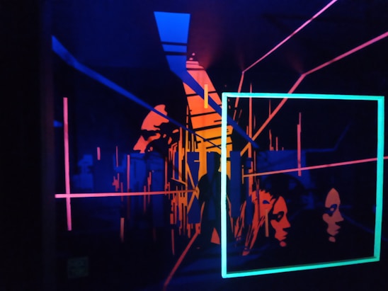 A vibrant, abstract artwork featuring neon outlines of human faces and geometric shapes. The composition includes intersecting lines and rectangles, creating a sense of depth and dimension against a dark background.