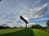 A large metal sculpture of an angel stands on a grassy hill. The sun is positioned directly behind the sculpture, creating a dramatic silhouette effect. The sky is filled with scattered clouds, and the foreground features green grass with a few people walking around the sculpture.