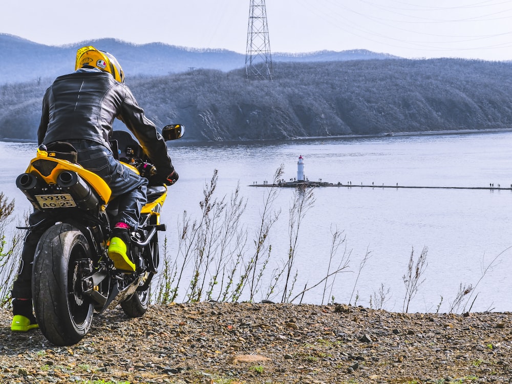 Homme en veste noire conduisant une moto jaune et noire près d’un plan d’eau pendant la journée