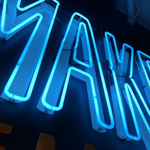 Bright neon letters in blue are illuminated against a dark background, creating a striking contrast. The neon tubes form bold and angular shapes, and are mounted on a wall with visible wiring connections.