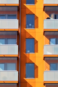 An apartment building facade with a striking orange color, featuring multiple windows and balconies. The balconies are lined with frosted glass railings, and the shadows cast by the balconies create geometric patterns on the orange walls.