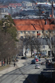 An urban setting featuring multiple stories of residential buildings with red roofs in the background and a busy street in the foreground. Cars populate the street, and a few pedestrians are visible walking on the sidewalks. Trees without leaves line the street, indicating a possible early spring or late autumn setting. The scene projects a sense of city life with a mixture of buildings, vehicles, and nature.