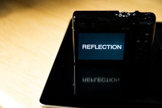 A digital camera displaying the word 'REFLECTION' on its screen, positioned on a shiny surface that reflects the camera and its display, under bright lighting conditions.