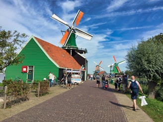 Several traditional Dutch windmills with green exteriors and red-tile roofs line a pedestrian pathway, under a bright blue sky with scattered clouds. People are walking and cycling along the path, enjoying the sunny day. Tall trees and bushes frame the scene, adding a touch of nature to the setting.