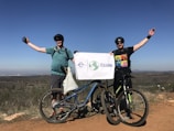 Two people are standing on a dirt path with bicycles in front of them, holding a sign that reads 'The Great Global Cleanup.' They are dressed in casual biking attire, wearing helmets and sunglasses. The landscape in the background shows open fields under a clear blue sky.