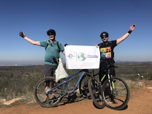 Two people are standing on a dirt path with bicycles in front of them, holding a sign that reads 'The Great Global Cleanup.' They are dressed in casual biking attire, wearing helmets and sunglasses. The landscape in the background shows open fields under a clear blue sky.