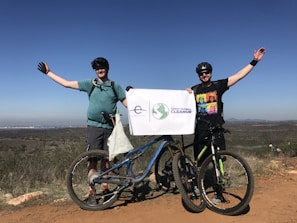 Two people are standing on a dirt path with bicycles in front of them, holding a sign that reads 'The Great Global Cleanup.' They are dressed in casual biking attire, wearing helmets and sunglasses. The landscape in the background shows open fields under a clear blue sky.
