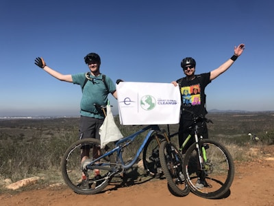 Two people are standing on a dirt path with bicycles in front of them, holding a sign that reads 'The Great Global Cleanup.' They are dressed in casual biking attire, wearing helmets and sunglasses. The landscape in the background shows open fields under a clear blue sky.