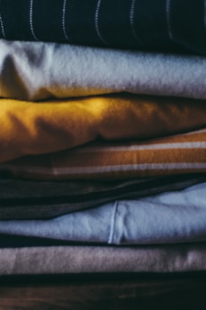 A stack of folded clothes in various colors including yellow, blue, and grey, arranged neatly one on top of another. The textures of the fabric are visible, with some items appearing made of cotton or similar materials.