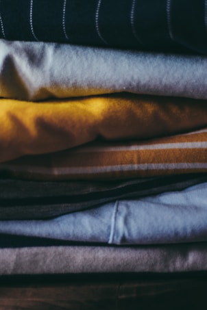 A stack of folded clothes in various colors including yellow, blue, and grey, arranged neatly one on top of another. The textures of the fabric are visible, with some items appearing made of cotton or similar materials.