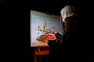 A person wearing a headscarf is painting a cityscape on a canvas. The scene includes tall towers and buildings with a variety of colors being mixed on a palette.