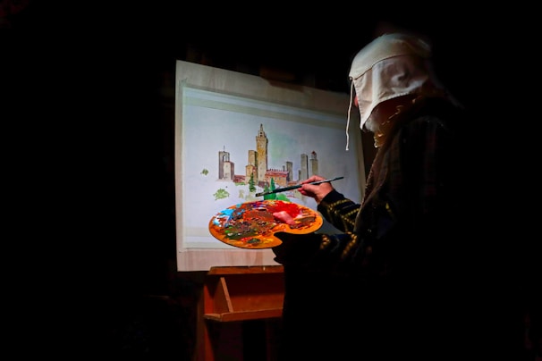 A person wearing a headscarf is painting a cityscape on a canvas. The scene includes tall towers and buildings with a variety of colors being mixed on a palette.