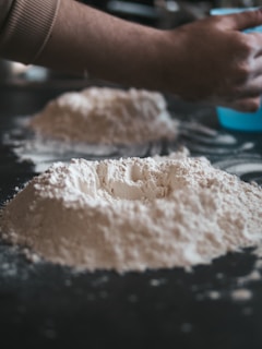 A close-up view of mounds of flour on a dark surface, with a hand reaching towards it, suggesting the preparation of dough.