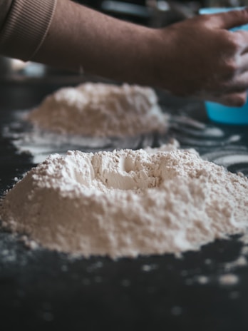 A close-up view of mounds of flour on a dark surface, with a hand reaching towards it, suggesting the preparation of dough.