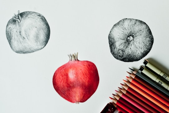 A drawing of three pomegranates featuring two sketched in black and white and one in color. The colored pomegranate is depicted in the center, with shades of red and hints of pink. To the right, various shades of red and pink colored pencils are neatly arranged along with two pens, suggesting an artistic workspace.