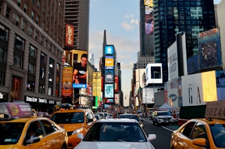 A bustling urban scene featuring a busy street lined with tall buildings covered in large advertisements and digital billboards. Yellow taxis are prominently visible in the foreground, with a mix of cars and pedestrians filling the area. The vibe is energetic and dynamic, indicative of a major cityscape.