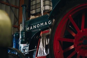 Vintage tractor with a turquoise body and large red spoked wheel, situated indoors. The name 'Hanomag' is prominently displayed above the wheel.