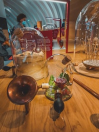 An interior space with a modern, curvilinear ceiling design. On a wooden table, there are glass domes containing items like raspberry jam, leaves, cork stoppers, and baguettes in burlap. A device with a brass trumpet-like horn is attached to the dome, suggesting it's an interactive exhibit, possibly at a museum or an exhibition. In the background, people are observing other displays, some wearing masks.