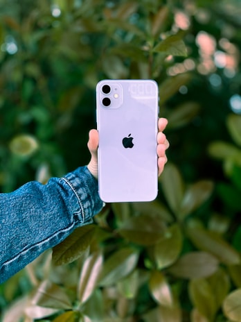 A hand holding a white smartphone with a dual-camera setup and the Apple logo on the back, set against a blurred background of green foliage. The hand is wearing a denim jacket.