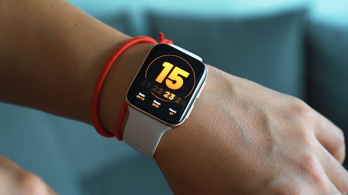 A smartwatch with a white band is displayed on a person's wrist. The watch screen shows a large number 15, with additional information including the date 09/12, steps 3995, and heart rate 107 bpm. The watch is worn above a red string bracelet on the wrist.