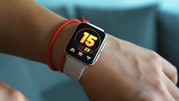 A smartwatch with a white band is displayed on a person's wrist. The watch screen shows a large number 15, with additional information including the date 09/12, steps 3995, and heart rate 107 bpm. The watch is worn above a red string bracelet on the wrist.