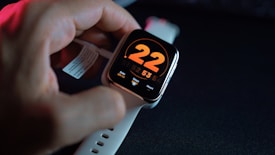 A smartwatch with a white strap is placed on a dark surface. The screen displays a large orange number 22, with smaller digital indicators below for date, steps, and heart rate. A hand is adjusting or holding the watch, and there's a paper tag attached to the strap.