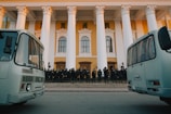 A group of uniformed individuals in protective gear, likely police or security personnel, stand in formation on the steps of a large building with classical architecture featuring tall white columns and a yellow facade. Two buses are parked prominently in the foreground.